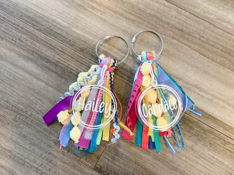 May include: Two clear acrylic keychains with colorful tassels. The keychains have the names "Hailey" and "Candace" written in white on them.