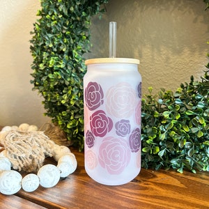May include: A frosted glass tumbler with a floral design in shades of pink and purple. The tumbler has a bamboo lid and a clear plastic straw.