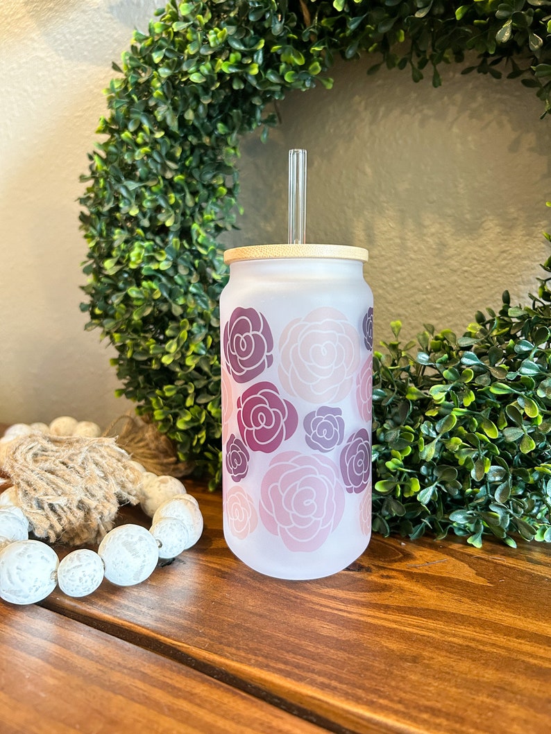 May include: A frosted glass tumbler with a floral design in shades of pink and purple. The tumbler has a bamboo lid and a clear plastic straw.