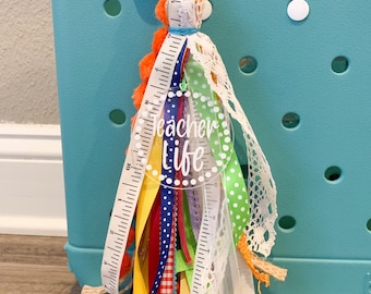 May include: A colorful teacher life tassel keychain with a clear acrylic charm that says "Teacher Life". The tassel is made of various ribbons, lace, and a ruler.