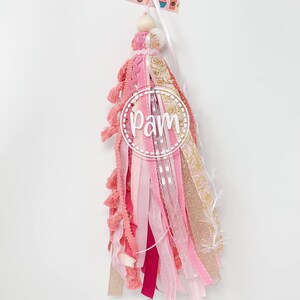 May include: Pink and gold tassel with a wooden bead and a white circle with the word "Pam" in it. The tassel is made of various ribbons and fabrics, including pink, gold, and white.