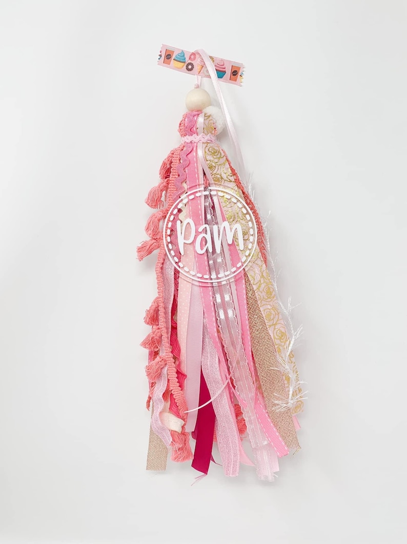 May include: Pink and gold tassel with a wooden bead and a white circle with the word "Pam" in it. The tassel is made of various ribbons and fabrics, including pink, gold, and white.