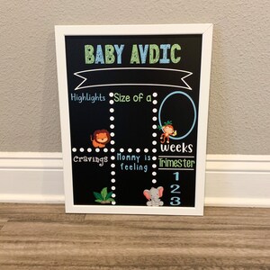 K&ouml;nnte beinhalten: Eine schwarze Tafel mit wei&szlig;em Rahmen, betitelt "BABY AVDIC". Die Tafel ist in Abschnitte mit den &Uuml;berschriften "Highlights", "Gr&ouml;&szlig;e eines", "Gel&uuml;ste", "Mama f&uuml;hlt sich", "Wochen" und "Trimester" unterteilt. Es gibt Illustrationen von einem L&ouml;wen, einem Affen und einem Elefanten.