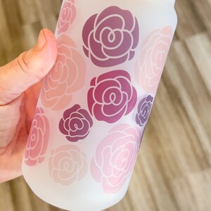 May include: A frosted glass water bottle with a bamboo lid and a clear plastic straw. The bottle has a floral design with pink and purple roses.