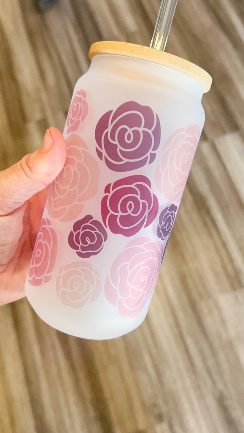 May include: A frosted glass water bottle with a bamboo lid and a clear plastic straw. The bottle has a floral design with pink and purple roses.