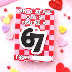 May include: A Valentine's Day card with a red and pink checkered background. The card reads "ON A SCALE OF 1-5, YOU'RE A 67 FROM," with a black and white "67" attached. Scattered around are heart-shaped candies and decorations.