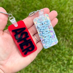 May include: Two rectangular keychains are displayed. One is red with the name "ASHER" in black, the other is clear with blue glitter and the name "ABIGAIL" in light blue. Both have silver key rings.