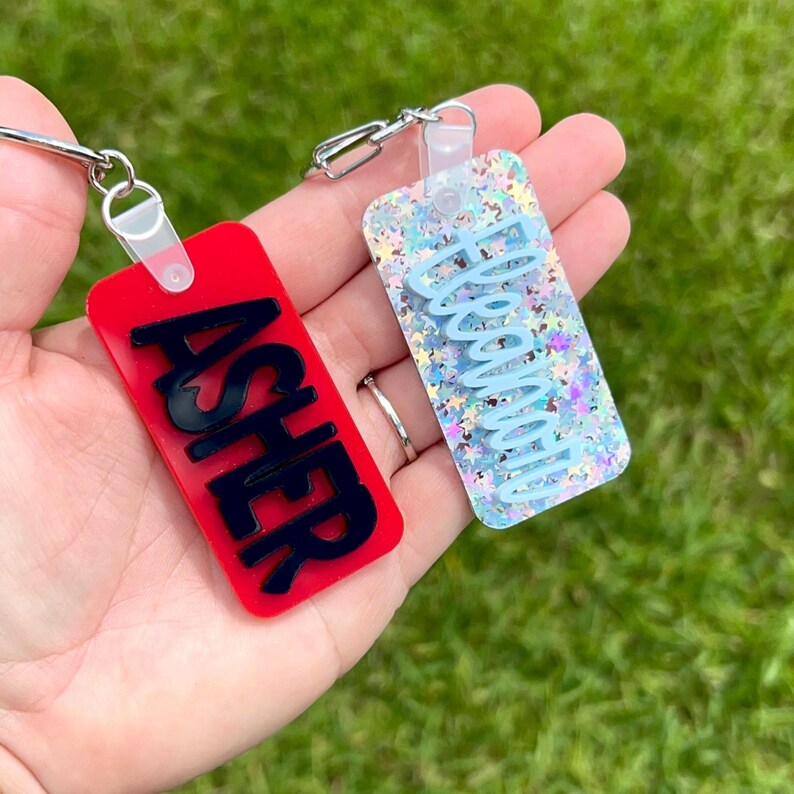 May include: Two rectangular keychains are displayed. One is red with the name "ASHER" in black, the other is clear with blue glitter and the name "ABIGAIL" in light blue. Both have silver key rings.