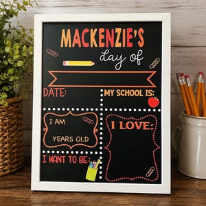 May include: A rectangular chalkboard sign in a white frame with the text MACKENZIE'S day of, DATE, MY SCHOOL IS, I AM YEARS OLD, I LOVE, and I WANT TO BE. The board features colorful graphics of a pencil, paper clips, and a red apple.