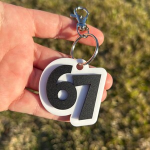May include: A white keychain with the number 67 in black. The keychain has a silver clasp and a metal ring. The background is blurred green grass.