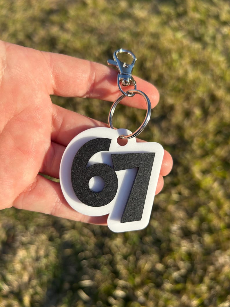 May include: A white keychain with the number 67 in black. The keychain has a silver clasp and a metal ring. The background is blurred green grass.