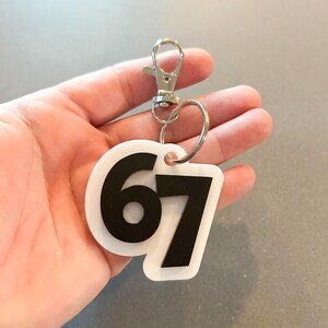 May include: White and black keychain with the number 67. The keychain has a silver clasp and a silver ring. The number 67 is in a bold, black font on a white background. The keychain is held in a hand.