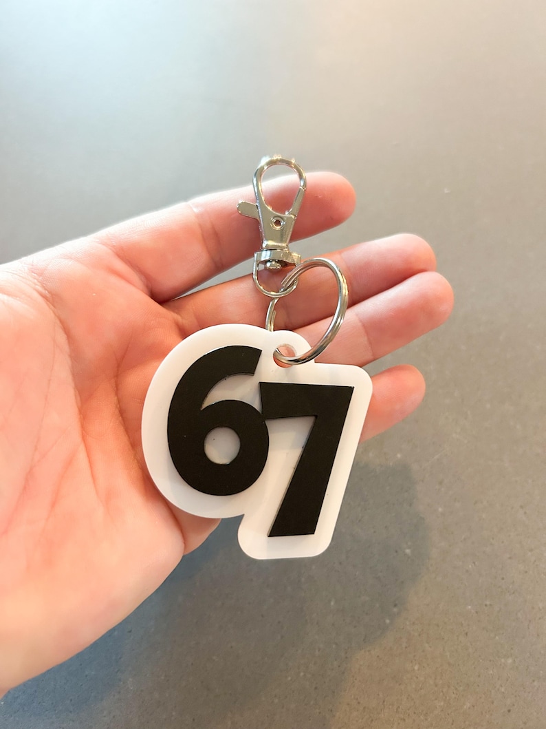 May include: White and black keychain with the number 67. The keychain has a silver clasp and a silver ring. The number 67 is in a bold, black font on a white background. The keychain is held in a hand.