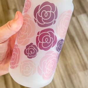 May include: A frosted glass reusable water bottle with a bamboo lid and a straw. The bottle has a pink and purple floral design.