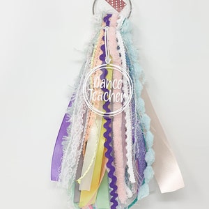 May include: A colorful key chain with a variety of ribbons, laces, and trims. The key chain has a metal ring and a brown polka dot ribbon. The key chain has a white circle with the text "Dance Teacher" in black.
