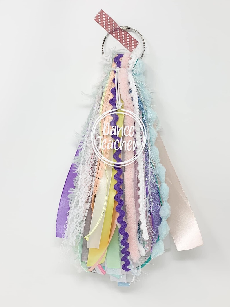 May include: A colorful key chain with a variety of ribbons, laces, and trims. The key chain has a metal ring and a brown polka dot ribbon. The key chain has a white circle with the text "Dance Teacher" in black.