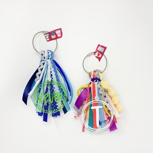 May include: Two colorful keychains with a circle of ribbon and a clear acrylic charm with a name or initial. The keychain on the left has a blue, white, and silver charm with the name "Kevin" on it. The keychain on the right has a red, pink, yellow, and green charm with the initial "T" on it.
