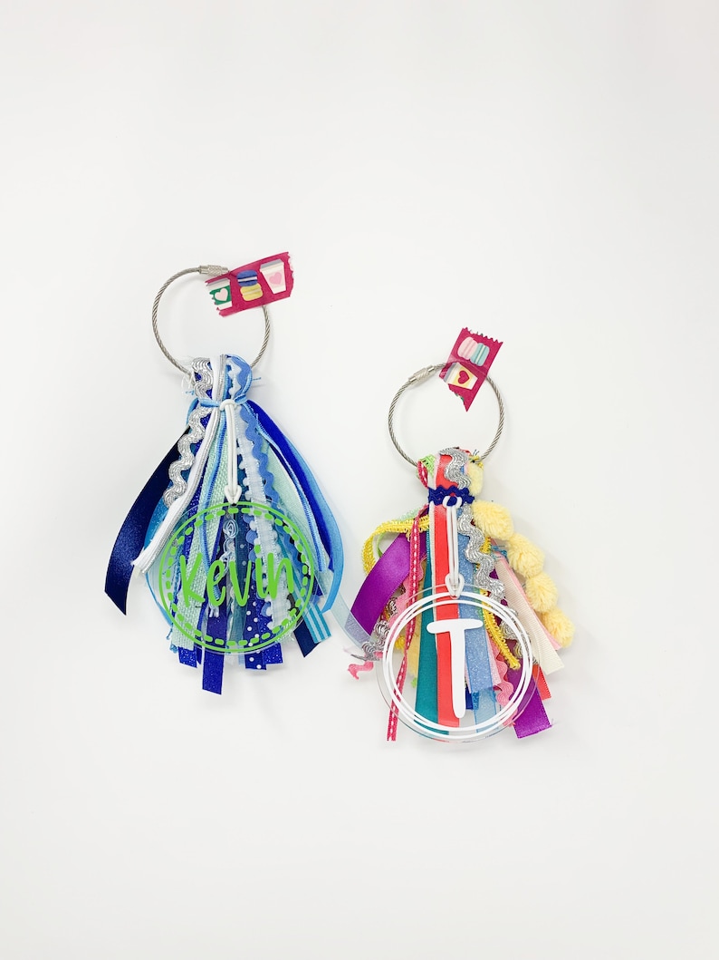 May include: Two colorful keychains with a circle of ribbon and a clear acrylic charm with a name or initial. The keychain on the left has a blue, white, and silver charm with the name "Kevin" on it. The keychain on the right has a red, pink, yellow, and green charm with the initial "T" on it.