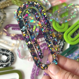 May include: A large, glitter-filled paperclip in a hand, with a black base and colorful iridescent glitter. Other paperclips in various colors and glitter patterns are in the background, including gold, pink, green, and purple.