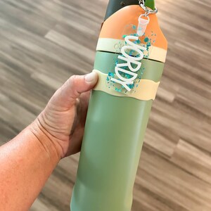 May include: A green and orange water bottle with a green handle and a clear tag that says "WORDY" in white letters. The tag has blue glitter inside. The bottle is held by a person.