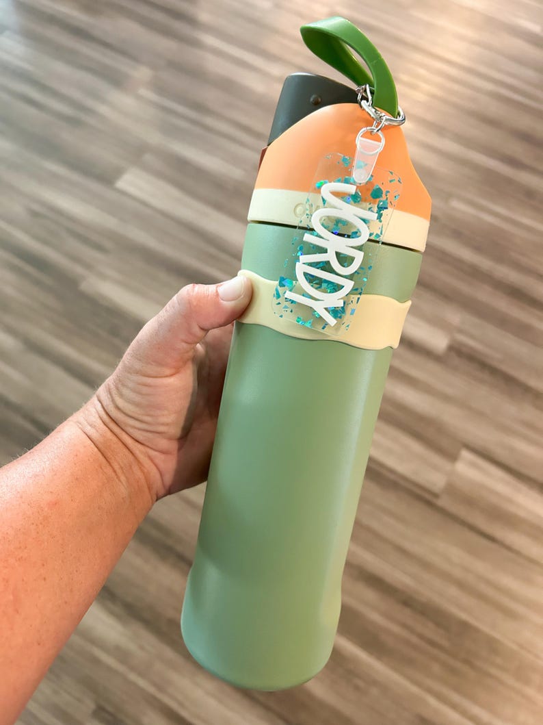 May include: A green and orange water bottle with a green handle and a clear tag that says "WORDY" in white letters. The tag has blue glitter inside. The bottle is held by a person.