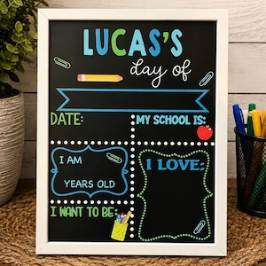 May include: A white-framed chalkboard sign featuring the text "LUCAS'S day of" with colorful graphics including a pencil, apple, and paper clips. The board has designated blank sections for date, school, age, interests, and future career goals.