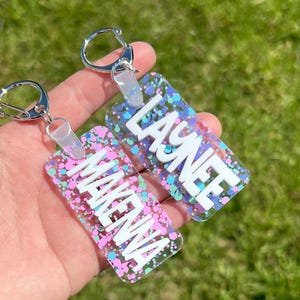May include: Two rectangular keychains with silver key rings. One keychain has the name "MAKENA" in white block letters, and the other has "LAYNEE". Both keychains have a clear background with pink, blue, and green glitter.