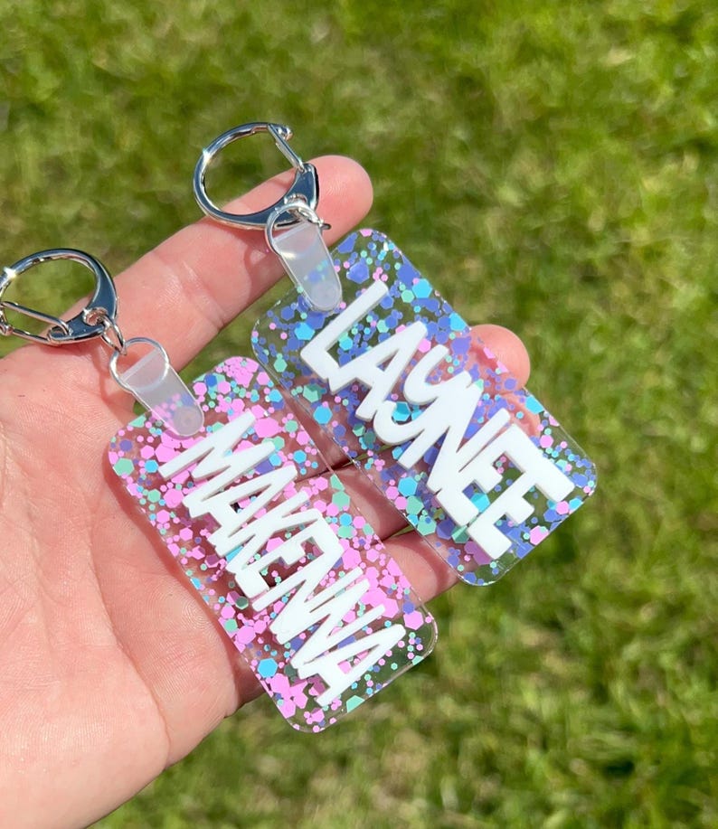 May include: Two rectangular keychains with silver key rings. One keychain has the name "MAKENA" in white block letters, and the other has "LAYNEE". Both keychains have a clear background with pink, blue, and green glitter.