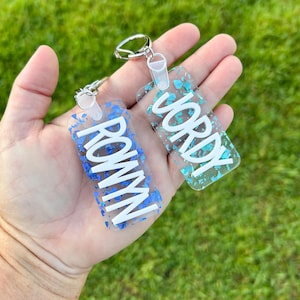May include: Two rectangular keychains with silver hardware. One keychain has the name "ROWYN" in white letters on a blue background with blue glitter. The other keychain has the name "JORDY" in white letters on a teal background with teal glitter.