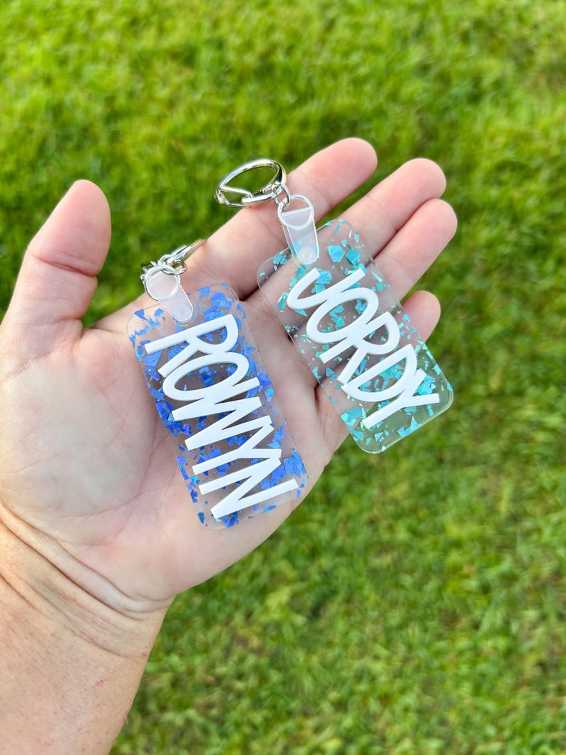 May include: Two rectangular keychains with silver hardware. One keychain has the name "ROWYN" in white letters on a blue background with blue glitter. The other keychain has the name "JORDY" in white letters on a teal background with teal glitter.