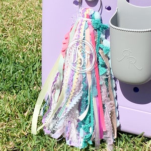 K&ouml;nnte beinhalten: Ein bunter Quasten-Schl&uuml;sselanh&auml;nger mit einem klaren Acryl-Anh&auml;nger mit dem Monogramm "DSF". Der Schl&uuml;sselanh&auml;nger ist an einer lila K&uuml;hltasche befestigt.