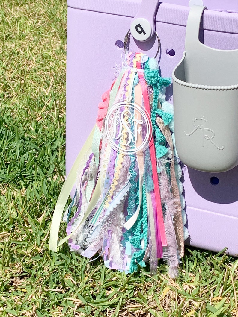 K&ouml;nnte beinhalten: Ein bunter Quasten-Schl&uuml;sselanh&auml;nger mit einem klaren Acryl-Anh&auml;nger mit dem Monogramm "DSF". Der Schl&uuml;sselanh&auml;nger ist an einer lila K&uuml;hltasche befestigt.