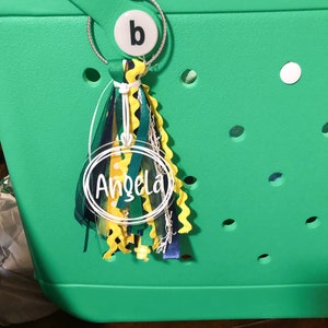 May include: A green tote bag with a clear round keychain that says "Angela" in white lettering. The keychain is attached to a silver ring with a white circle that has the letter "b" on it. The keychain also has a multi-colored tassel.