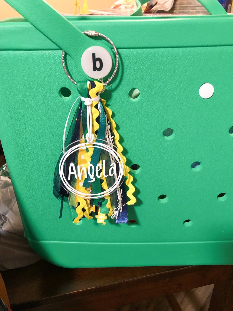 May include: A green tote bag with a clear round keychain that says "Angela" in white lettering. The keychain is attached to a silver ring with a white circle that has the letter "b" on it. The keychain also has a multi-colored tassel.