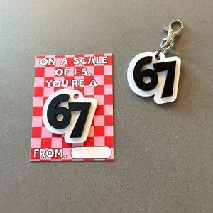 May include: A white and black keychain and card set. The keychain features the number 67 in black on a white background with a silver clasp. The card has a red and pink checkered pattern with the text "ON A SCALE OF I-5, YOU'RE A 67 FROM."