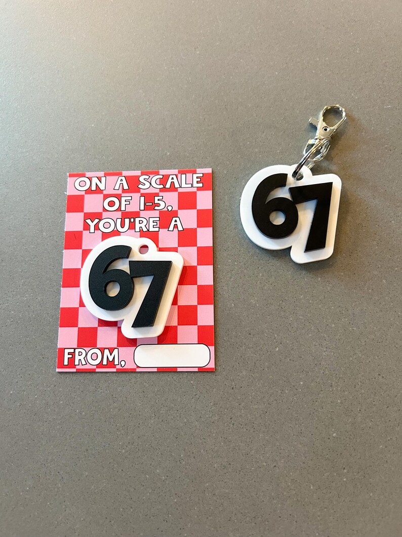 May include: A white and black keychain and card set. The keychain features the number 67 in black on a white background with a silver clasp. The card has a red and pink checkered pattern with the text "ON A SCALE OF I-5, YOU'RE A 67 FROM."