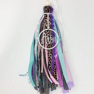 May include: A white tassel with a variety of ribbons in shades of pink, purple, teal, and gray. The tassel has a white circle with the initials "LW" in black lettering.
