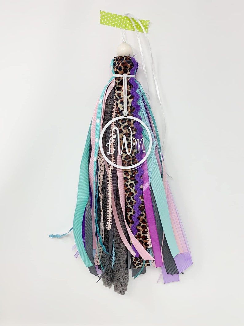May include: A white tassel with a variety of ribbons in shades of pink, purple, teal, and gray. The tassel has a white circle with the initials "LW" in black lettering.