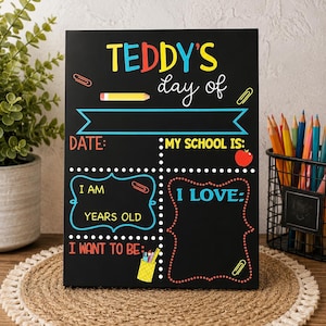 May include: A black rectangular school-themed sign with colorful text and graphics. It features the heading TEDDY'S day of, with fillable sections for date, school, age, interests, and future career goals, decorated with pencils, paperclips, and an apple.