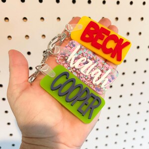 May include: Three personalized keychains with names: "BECK" in red on yellow, "Keriah" in white on a glittery background, and "COOPER" in dark blue on green. The keychains are attached to a silver clasp.