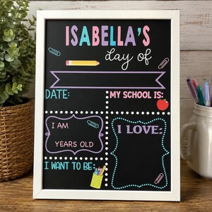 May include: A rectangular chalkboard sign in a white frame featuring colorful text and graphics. The board reads ISABELLA'S day of, DATE, MY SCHOOL IS, I AM YEARS OLD, I LOVE, and I WANT TO BE, with illustrations of a pencil, apple, and pen holder.