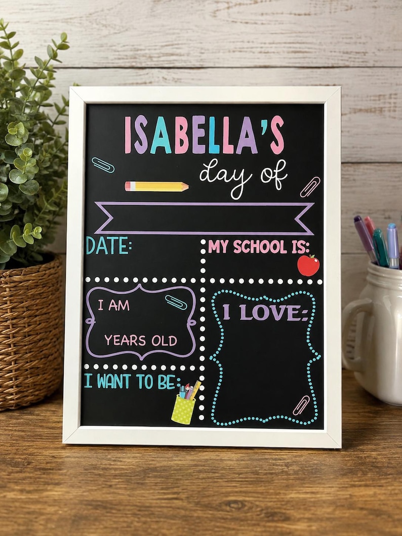 May include: A rectangular chalkboard sign in a white frame featuring colorful text and graphics. The board reads ISABELLA'S day of, DATE, MY SCHOOL IS, I AM YEARS OLD, I LOVE, and I WANT TO BE, with illustrations of a pencil, apple, and pen holder.