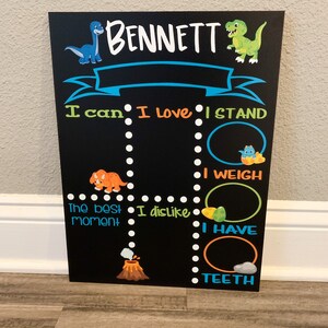 Puede incluir: Una pizarra negra con tem&aacute;tica de dinosaurios. Tiene un dinosaurio azul, un dinosaurio verde y un volc&aacute;n. La pizarra tiene el nombre "BENNETT" en la parte superior y dice "Puedo", "Me encanta", "Me pongo de pie", "Peso", "El mejor momento", "No me gusta", "Tengo", y "Dientes".