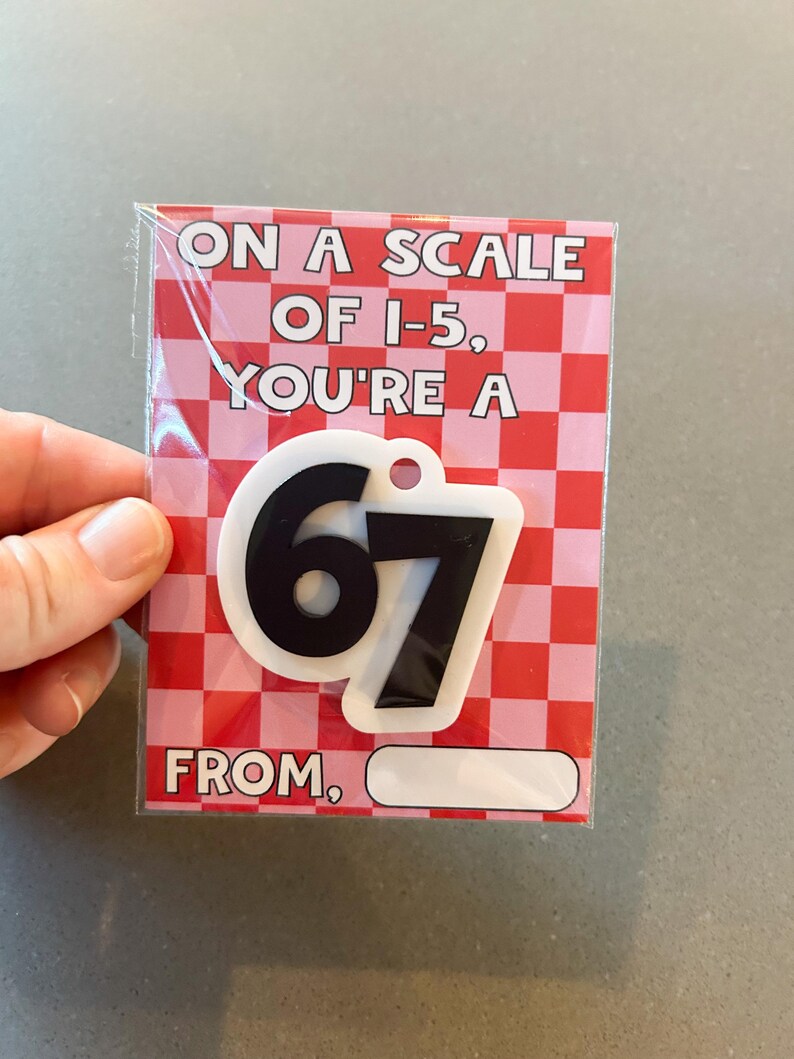 May include: A greeting card with a pink and red checkered background. The card reads "ON A SCALE OF 1-5, YOU'RE A 67" with a black and white keychain of the number 67. The bottom of the card has a blank space that says "FROM,"