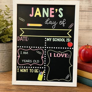 May include: A white-framed chalkboard sign with colorful text reading JANE'S day of, DATE:, MY SCHOOL IS:, I AM YEARS OLD, I LOVE:, and I WANT TO BE:. The board features illustrations of a yellow pencil, a red apple, and a green pencil holder.