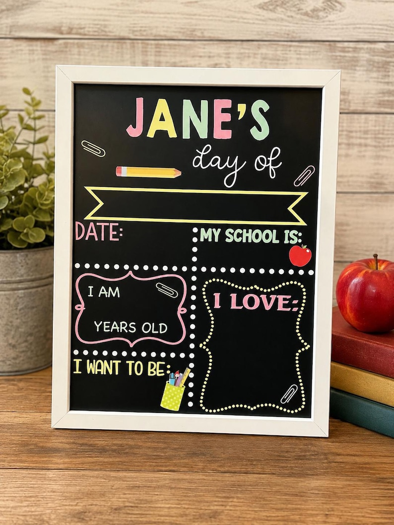 May include: A white-framed chalkboard sign with colorful text reading JANE'S day of, DATE:, MY SCHOOL IS:, I AM YEARS OLD, I LOVE:, and I WANT TO BE:. The board features illustrations of a yellow pencil, a red apple, and a green pencil holder.