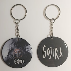 May include: Two black and silver keychains with round metal tags. One tag features a black and white image of a creature with the word "GOJIRA" printed below. The other tag has the word "GOJIRA" printed in white on a black background.