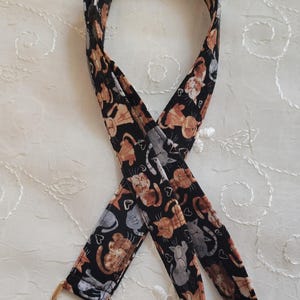 May include: A fabric lanyard featuring a repeating pattern of orange, brown, and gray cats interspersed with small white hearts on a black background. The strap is finished with a gold-toned metal swivel lobster clasp at the bottom end.