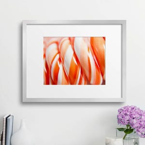 May include: Close-up of a framed print featuring a cluster of orange and white swirled candy canes. The image is displayed on a white desk with a laptop, books, scissors, and a vase of purple flowers.