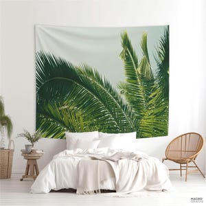 a bedroom with a large tapestry of palm leaves hanging on the wall above a bed. The bed is covered in white linens and has a wooden chair nearby. The room has a minimalist and modern design, with a white color scheme and natural elements like the palm leaves.